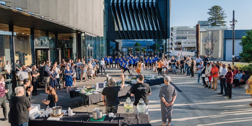 A group of guests attending the Amotai: Meet the Buyers event, gathered around a tables filled with local food, while a group of young students from the Tauranga Intermediate Kapa Haka group perform.